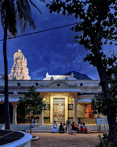 Floodlit gopuram and shrines of Sri Ramanasramam at dusk beneath Arunachala, with lit verandas and walkways after sunset